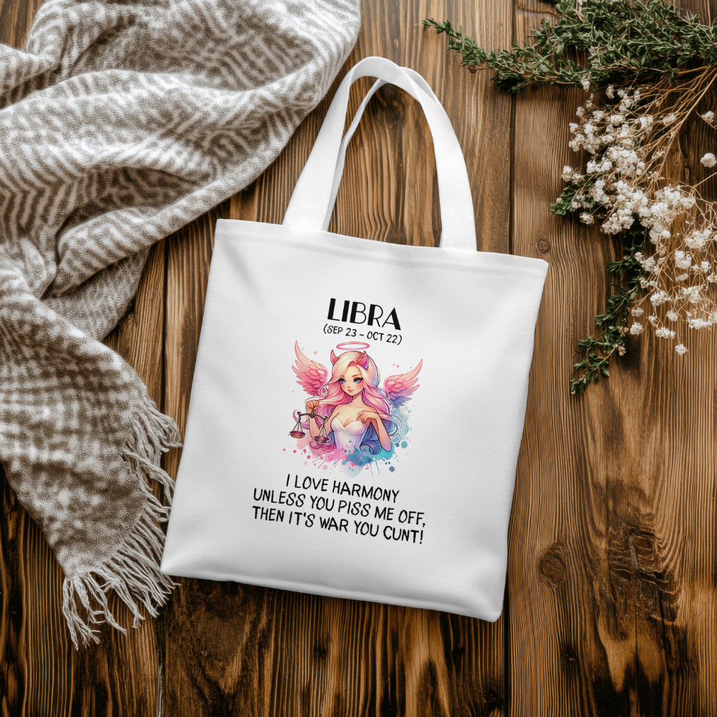 A white satin-look 100% polyester tote bag (38cm x 40cm) featuring a bold Libra-themed design. The artwork showcases a pink-haired angelic figure holding the Libra scales, with soft pastel wings and a dreamy colour palette. Below, the cheeky text reads: "I love harmony unless you piss me off, then it's war you cunt!" A sassy, funny astrology-themed tote bag, perfect for Libra star sign lovers.