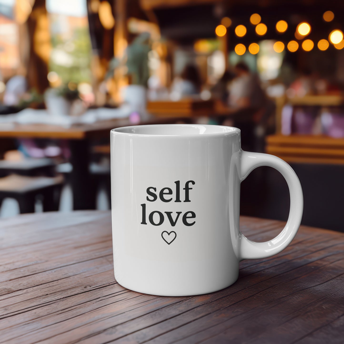 White 11oz ceramic mug with the words "self love" and a small black heart printed in minimalist black font on both sides. Shown in a cafe setting and in a hand for size reference.