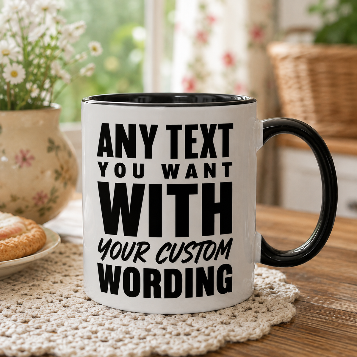 A white ceramic mug with a black handle and black interior, printed with the ANY TEXT YOU WANT WITH YOUR CUSTOM WORDING' in bold black lettering. The mug sits on a beige crocheted doily on a wooden table, with a blurred kitchen background showing a floral vase, flowers, and a wicker basket.