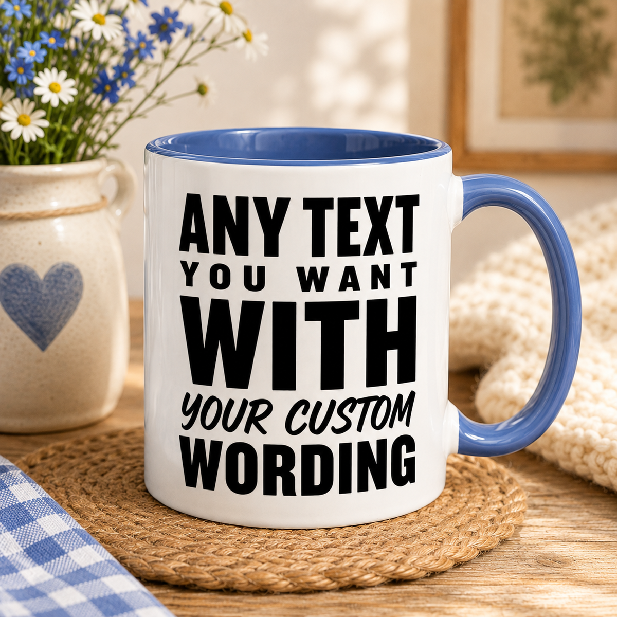 A white ceramic coffee mug with a blue interior and handle, printed with ANY TEXT YOU WANT WITH YOUR CUSTOM WORDING' in bold black letters. The mug sits on a woven placemat on a wooden table, surrounded by cozy lifestyle props including a blue and white gingham cloth, a cream knit blanket, and a small ceramic vase of daisies.