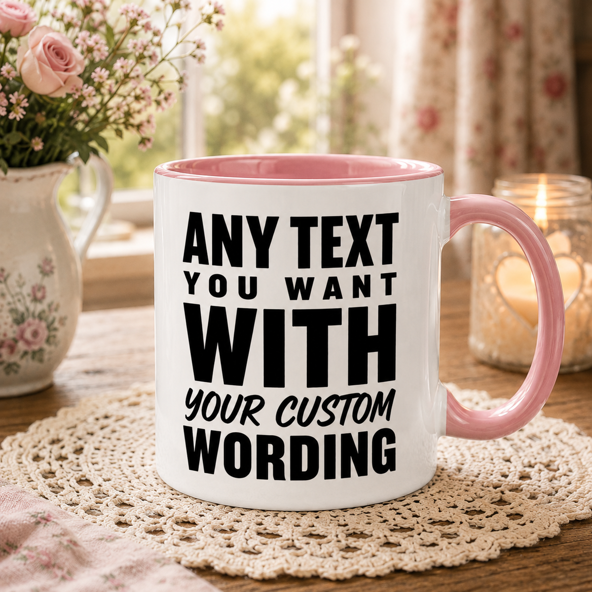 A white ceramic mug with a pink handle and interior, printed with ANY TEXT YOU WANT WITH YOUR CUSTOM WORDING' in black. The mug sits on a lace doily on a wooden table, with a blurred background showing a window, flowers, and a lit candle.