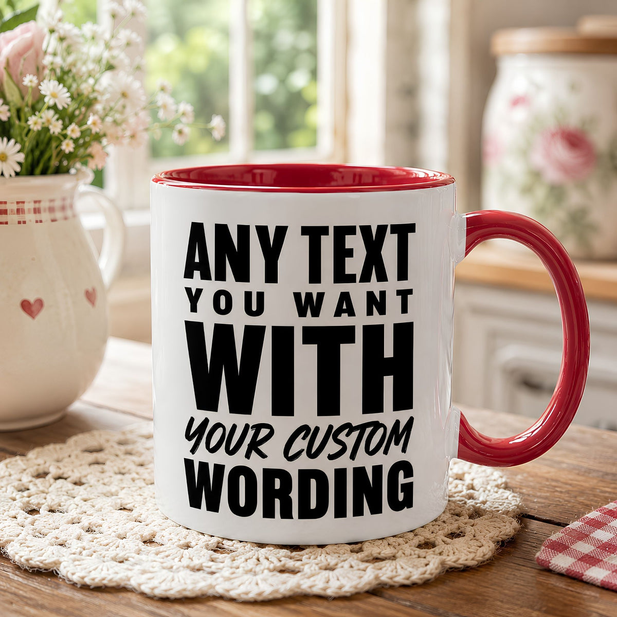 A white ceramic mug with a red handle and red interior, printed with the customizable text 'ANY TEXT YOU WANT WITH YOUR CUSTOM WORDING' in bold black lettering. The mug sits on a cream crocheted doily on a wooden table, with a blurred background showing a window, flowers, and a decorative jar.