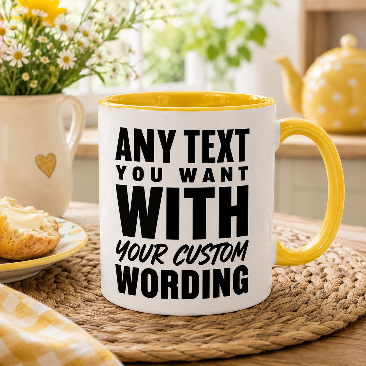 A white ceramic mug with a yellow interior and handle, printed with ANY TEXT YOU WANT WITH YOUR CUSTOM WORDING' in bold black lettering. The mug is placed on a woven placemat on a wooden table, with a blurred breakfast scene in the background featuring a teapot, flowers, and a plate of toast.