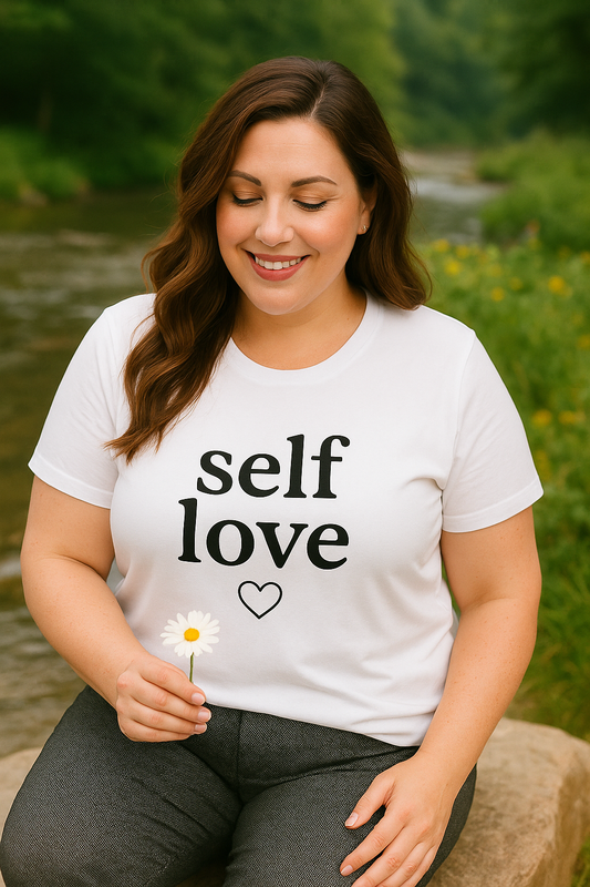 A smiling woman sits peacefully by a river holding a daisy, wearing a fitted white t-shirt with the phrase "self love" and a heart outline. This empowering image promotes body positivity, self-care, and mental health awareness, showcasing the soft cotton tee's comfortable fit and meaningful message.