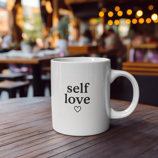 White 11oz ceramic mug with the words "self love" and a small black heart printed in minimalist black font on both sides. Shown in a cafe setting and in a hand for size reference.