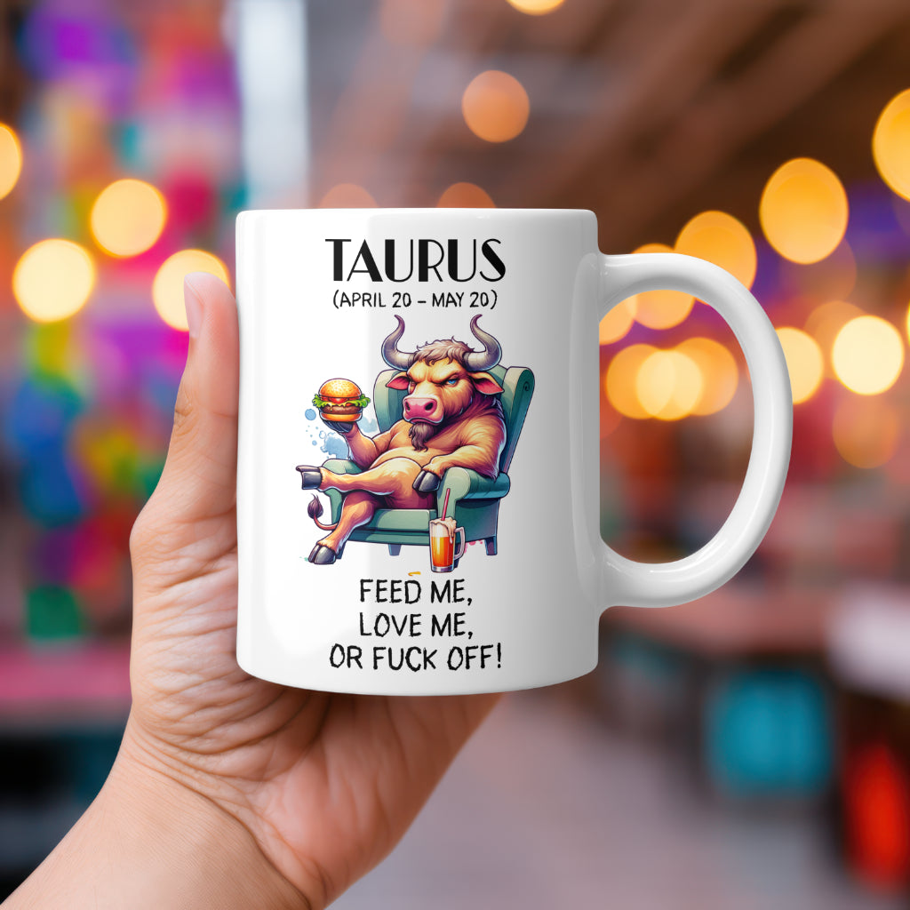 A white 11oz coffee mug featuring a bold Taurus-themed design. The artwork showcases a relaxed bull lounging in a green armchair, holding a juicy burger in one hand and resting near a drink on the side. The Taurus zodiac symbol is in the background. Below, the cheeky text reads: "Feed me, love me, or fuck off!" A hilarious, rude astrology gift for Taurus star sign lovers.