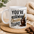 White mug with humorous text that says - You're about as useful as a chocolate teapot and teapot illustration on a wooden surface with chocolate and a plant.